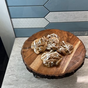 Puede incluir: Una bandeja de madera con borde natural contiene varias pralin&eacute;s de nueces pecanas rociadas con glaseado blanco. La bandeja se encuentra sobre una encimera de color claro, con un protector contra salpicaduras de azulejos azules y blancos estampados en el fondo.