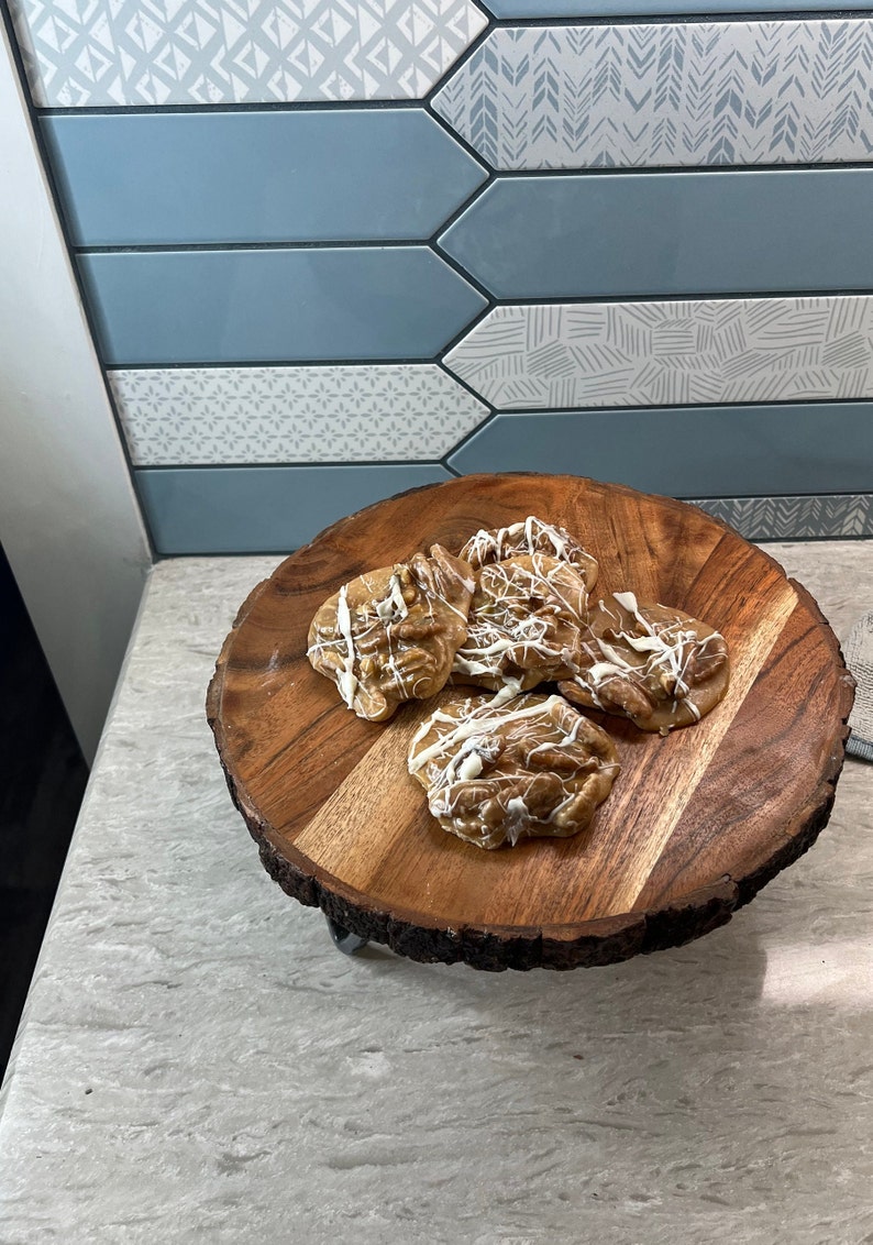 Puede incluir: Una bandeja de madera con borde natural contiene varias pralin&eacute;s de nueces pecanas rociadas con glaseado blanco. La bandeja se encuentra sobre una encimera de color claro, con un protector contra salpicaduras de azulejos azules y blancos estampados en el fondo.