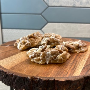 Puede incluir: Varios caramelos de nuez pecana rociados con glaseado blanco est&aacute;n dispuestos en una tabla redonda de madera con borde natural. Los caramelos son de color marr&oacute;n claro y tienen una superficie texturizada. La tabla tiene corteza marr&oacute;n oscuro.