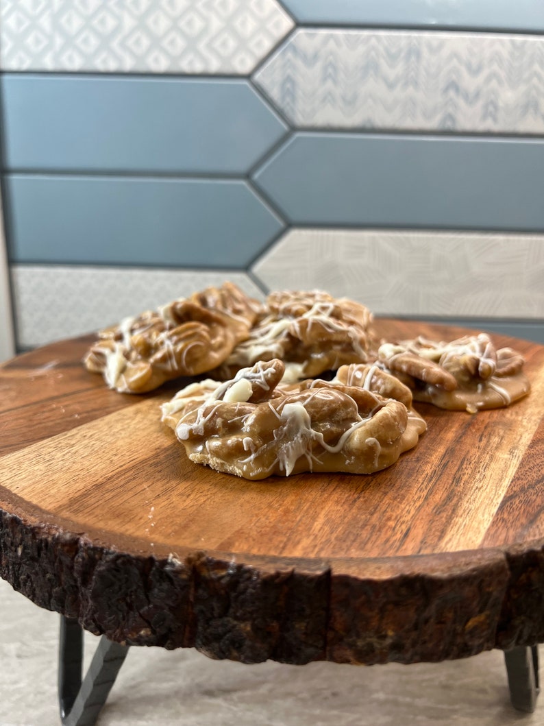 Puede incluir: Varios caramelos de nuez pecana rociados con glaseado blanco est&aacute;n dispuestos en una tabla redonda de madera con borde natural. Los caramelos son de color marr&oacute;n claro y tienen una superficie texturizada. La tabla tiene corteza marr&oacute;n oscuro.