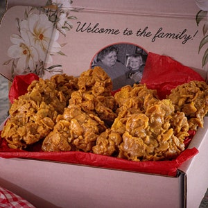 May include: A box of golden-brown, textured cookies, possibly a type of brittle or cluster, sits on a red paper liner. The box is decorated with floral accents and the text "Welcome to the family!" A photo of two people is inside the box.