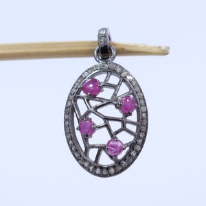 Op de afbeelding: Een zilveren hanger met een delicaat, ingewikkeld ontwerp met een ronde rand met een zwarte geoxideerde afwerking. De hanger is versierd met vier roze edelstenen en kleine, heldere diamanten.