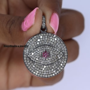 Puede incluir: Un colgante de plata con un diseño de ojo malvado redondo incrustado de diamantes negros. El ojo está acentuado con un pequeño rubí rojo en el centro.