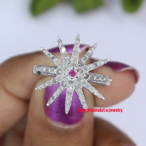 Puede incluir: Un anillo de plata con un diseño de estrella de explosión con una piedra roja en el centro y pequeños diamantes alrededor de la estrella. El anillo está en un dedo con esmalte de uñas morado.
