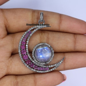 Pode incluir: Um pingente de prata em forma de lua crescente com uma grande pedra de lua redonda no centro. A lua é adornada com pequenos diamantes redondos e rubis cor-de-rosa.