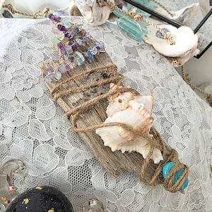 May include: A piece of weathered driftwood wrapped in brown twine, featuring a cluster of small purple and clear tumbled stones at the top, a large white seashell in the center, and a single smooth turquoise stone at the bottom end.