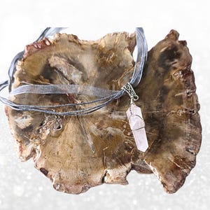 May include: A rose quartz crystal pendant necklace. The crystal is wrapped in silver wire and hangs from a silver ribbon and gray cord. The necklace is displayed on a brown, textured surface.