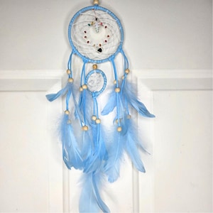 May include: A light blue dream catcher featuring a circular frame wrapped in matching cord, a white webbed center with small colorful beads, and a smaller secondary hoop. Light blue feathers and wooden beads hang from the bottom of the structure.