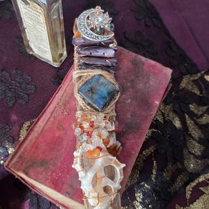 May include: A decorative wand featuring a silver-toned crescent moon and sun charm at the top, followed by dark purple rectangular stones, a central square blue labradorite, a cluster of small orange and clear crystals, and a white seashell at the base.