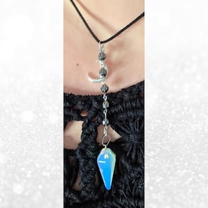 Puede incluir: Un collar con colgante que presenta un cordón negro, una luna creciente plateada y un péndulo de cristal azul. El collar tiene cuentas grises oscuras y un péndulo de cristal azul en forma de lágrima. El collar se usa sobre un fondo negro texturizado.