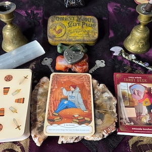 Puede incluir: Una colección de objetos místicos que incluyen una baraja de tarot, cartas, una lata vintage, un candado en forma de corazón, llaves y una varita de selenita. La carta del tarot presenta una ilustración de una persona con una sudadera con capucha. La lata dice "HONEST LABOR CUT PLUG".