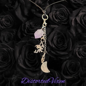 May include: A silver-toned charm necklace featuring a light purple amethyst stone, a dragon charm, and a crescent moon pendant. The necklace hangs against a backdrop of dark roses. The text "Distorted Vixen" is at the bottom.