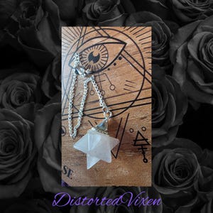 May include: A clear quartz crystal pendulum with a silver chain and cap hangs in front of a wooden surface with engraved symbols. The background features a dark floral pattern and the text "Distorted Vixen".