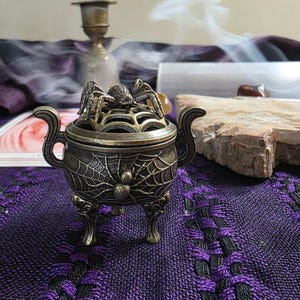 May include: A bronze-toned metal incense burner featuring a three-legged cauldron design with two side handles. The exterior is embossed with a spider and web pattern, and the removable lid is topped with a sculpted spider figure.