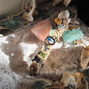 May include: A wooden wand wrapped in brown twine, decorated with small seashells, a silver-toned metal charm, and various pieces of translucent sea glass in shades of blue and green, featuring a small spiral shell attached to the bottom end.