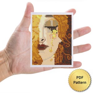 Puede incluir: Pequeña obra de arte de punto de cruz que representa un rostro estilizado en tonos marrones, dorados y beige. La pieza se sostiene en una mano. Un sello dorado en la esquina inferior derecha dice 'PDF Pattern'.
