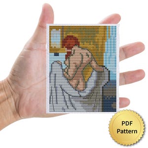 May include: A cross-stitch pattern featuring a nude figure with red hair, draped in a white cloth, against a backdrop of gold and blue. The image is held in a hand, with a gold PDF Pattern seal.