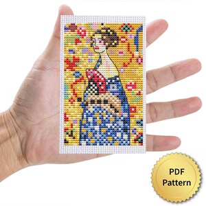 May include: A cross-stitch pattern held in a hand, featuring a woman in a blue dress with a floral background. The design has a yellow background with red, blue, and green floral accents. A gold-colored seal with the text "PDF Pattern" is in the lower right corner.