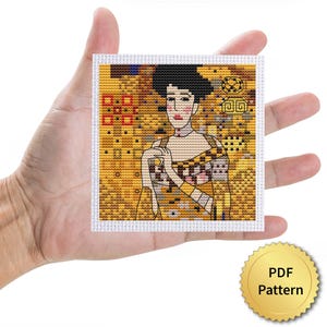 May include: A cross-stitch pattern featuring a woman in a yellow and gold dress with a black hairstyle. The artwork is held in a hand, and a gold-colored PDF pattern seal is in the lower right corner.