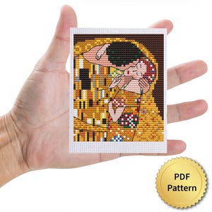 May include: A cross-stitch pattern of Gustav Klimt's "The Kiss" held in a hand. The artwork features gold, yellow, and brown hues with accents of green, red, and white. The image is a PDF pattern.