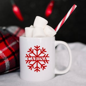May include: White ceramic mug with a red snowflake design and the name "CARINA" written inside the snowflake. The mug is filled with marshmallows and a red and white striped straw.
