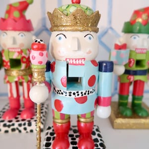 May include: Three decorative wooden nutcrackers. The central nutcracker is light blue with red polka dots, a gold crown, and red boots. The other two nutcrackers are red and green. Each stands on a black and white patterned base.