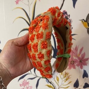 May include: A knotted headband with a brown base and orange checkered pattern. The headband is made of a soft, textured fabric.