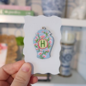 May include: A light blue decorative wooden ornament shaped like a vase, adorned with pink flowers and the letter "H" in a green frame. The ornament is held against a white, scalloped-edge card, suggesting a craft or home decor item.