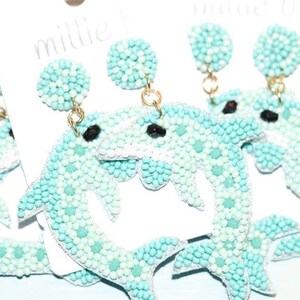 May include: A pair of blue and white beaded dolphin earrings. The dolphins are facing each other and are connected by a gold chain.