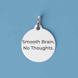 May include: A white, circular tag with a silver-colored ring at the top. The tag has the text "Smooth Brain. No Thoughts." in black, centered on the front. The background is a solid light blue.