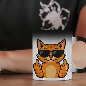 Puede incluir: Taza que cambia de color en blanco y negro con un gato de dibujos animados con gafas de sol y mostrando el dedo corazón. El gato es naranja con rayas negras. La taza tiene un asa negra.