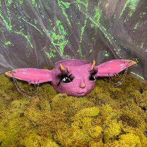 Könnte beinhalten: Ein skurriler, lavendelfarbener Pflanztopf in Form eines Kreaturengesichts mit großen, rosa Ohren und Wimpern. Der Topf ist mit kleinen Hörnern und silbernen Ketten verziert. Er ruht auf grünem Moos, mit einem schimmernden Hintergrund.