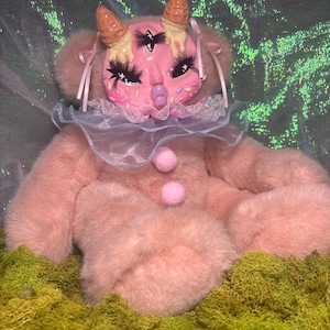 May include: A pink plush doll with a whimsical face, adorned with ice cream cone horns and a ruffled collar. The doll has long eyelashes and a three-eyed design. The doll is set against a green and iridescent background.