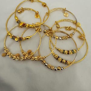 May include: A set of nine gold bangles with various designs, including beaded, twisted, and plain styles. Some bangles have small charms, such as hearts and bells.