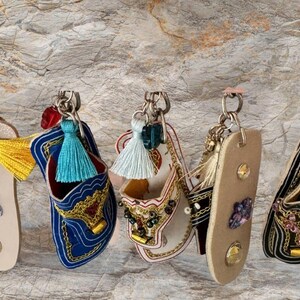 May include: Five colorful leather slipper keychains with beaded embellishments and tassels. The keychains are hanging from silver key rings.