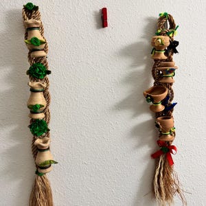 以下が含まれることがあります： 小さな陶器の鉢と緑色のアクセントが付いた、2つの装飾的なロープオーナメント。一方のオーナメントには赤いリボン、もう一方には緑色の布の花が付いています。赤い洗濯バサミがオーナメントの間の壁にあります。