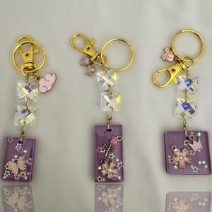 May include: Three purple resin keychain charms with gold key rings and iridescent crystal accents. Each charm features a different design: a pink flower with a green stem, a pink flower with a gold crown, and a pink butterfly with stars.