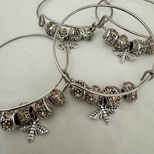 May include: Silver-toned bangle bracelets with decorative beads and bee charms. The bracelets feature a variety of round, textured beads in shades of green and peach. Each bracelet has a small, detailed bee charm.