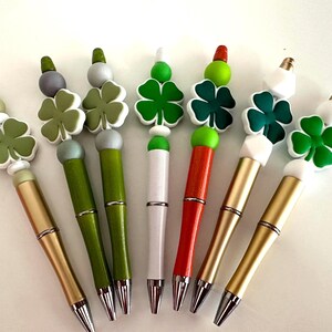 May include: Seven pens with green four-leaf clover charms. The pens have different colored barrels: gold, green, white, and orange.