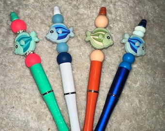 May include: Four colorful pens with fish-shaped toppers. The pens are green, white, orange, and blue. Each fish topper has a different color scheme and bead accents.