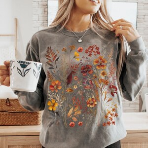 May include: Gray sweatshirt with a floral design in shades of orange, yellow, and red. The sweatshirt is paired with black biker shorts. A white mug with a blue floral design is held in the hand.