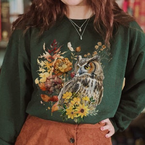 May include: A dark green sweatshirt with a graphic print of an owl surrounded by fall foliage. The owl is brown and gray with yellow eyes. The foliage is a mix of yellow, orange, and brown.