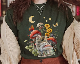 May include: A dark green t-shirt with a whimsical illustration of a cat sitting amongst mushrooms and flowers. The design features a crescent moon, stars, and butterflies.