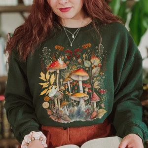 Pode incluir: Uma mulher vestindo uma camisa de frio verde com uma estampa de cogumelos e o texto "Shroom Queens". Ela est&aacute; segurando um livro em suas m&atilde;os.