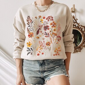 May include: A beige sweatshirt with a floral print featuring various shades of orange, yellow, and red. The flowers are arranged in a cluster on the front of the sweatshirt.