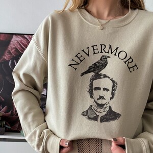 May include: A beige sweatshirt with a black graphic print of Edgar Allan Poe with a raven perched on his head. The text "NEVERMORE" is printed above the graphic.