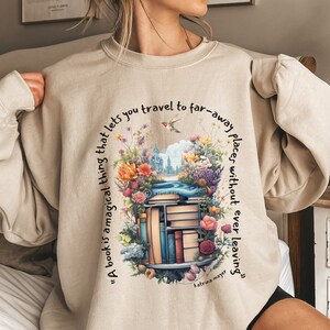 K&ouml;nnte beinhalten: Ein beiger Pullover mit einem grafischen Druck eines B&uuml;cherstapels, umgeben von Blumen und einem Zitat, das lautet: "Ein Buch ist eine magische Sache, die es dir erm&ouml;glicht, an ferne Orte zu reisen, ohne jemals wegzugehen" von Katrina Mayer.