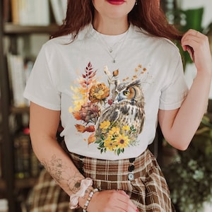 May include: White t-shirt with a graphic print of an owl surrounded by fall foliage. The owl is brown and gray with yellow eyes. The foliage is in shades of brown, yellow, and orange.