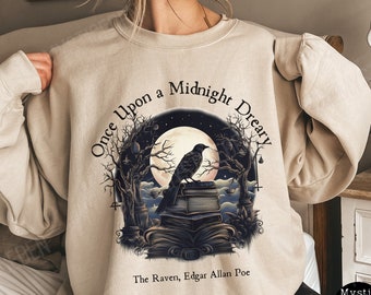 May include: A beige sweatshirt featuring a graphic print of a raven perched on a stack of books beneath a full moon. The text reads "Once Upon a Midnight Dreary" and "The Raven, Edgar Allan Poe".