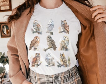 May include: White t-shirt with a watercolor design of nine owls in various poses. The owls are brown, white, and gray with black eyes. The shirt has a crew neck and short sleeves.